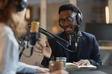 Medium shot of young Black man in eyeglasses and suit answering questions of unrecognizable...