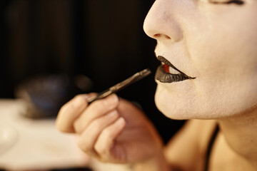 Side view close up of unrecognizable mime performer doing makeup backstage and painting black lips,...