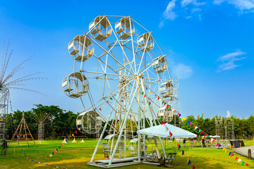 Beautiful scenic with white ferris wheel decorated with lights and small flags for fun and enjoyment of festival or carnival stand on green lawn. Outdoor theme park with beautiful nature as background