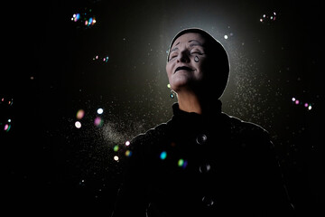 Minimal portrait of smiling mime artist playing with soap bubbles against black background, copy...