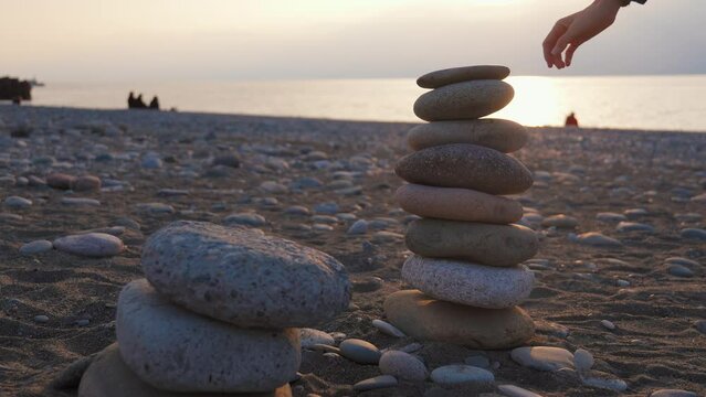 Spiritually relaxed woman putting stone on pyramid of cobblestones on pebble beach by sea at sunset. Yoga female peace of mind relax meditation harmony life balance tranquility equilibrium pyramid
