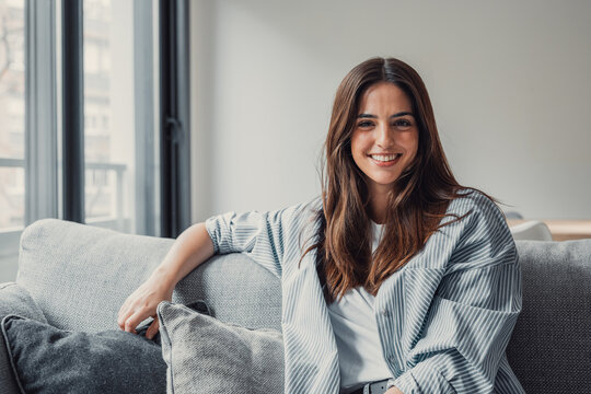 Happy attractive young adult woman girl sitting on sofa looking at camera in modern cozy home, smiling single 20s lady beautiful face relaxing on couch alone enjoy wellbeing posing for portrait.