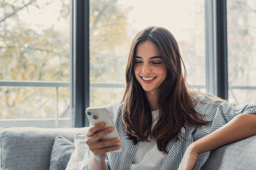 Happy pleasant millennial woman relaxing on comfortable couch, holding smartphone in hands. Smiling...