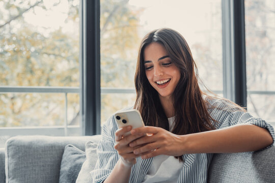 Happy pleasant millennial woman relaxing on comfortable couch, holding smartphone in hands. Smiling young lady chatting in social networks, watching funny videos, using mobile applications at home..