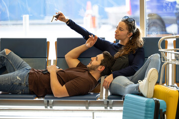 couple sitting on airport benches while taking a selfie waiting for their flight to depart