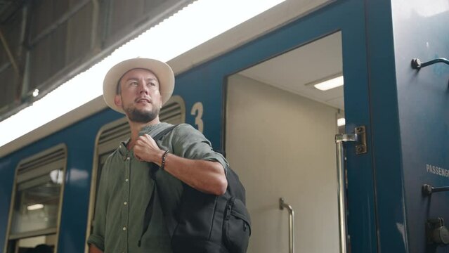 Asian male traveler steps off train, explores station platform. Adventure seeker wears, carries backpack. Rail journey, tourist exits carriage, begins city exploration. Summer, wanderlust, transit.