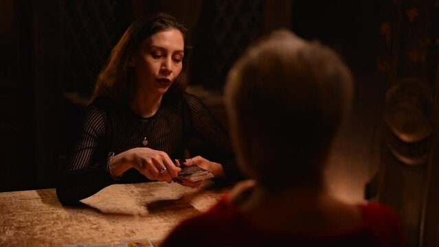 Unrecognizable adult female client getting forecasting future during divination session with tarot cards at dark esoteric room. Fortune teller tarot reader placing cards in order as mystic rite.