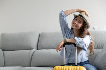 happy woman sitting on the sofa with her hat on and hand suitcase ready to go on summer vacation