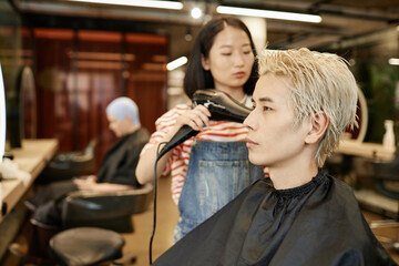 Side view portrait of young Asian man in beauty salon with hairstylist drying bleached hair, copy space