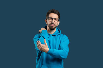 A young man wearing glasses and blue hoodie stands against plain blue backdrop, touching elbow with...