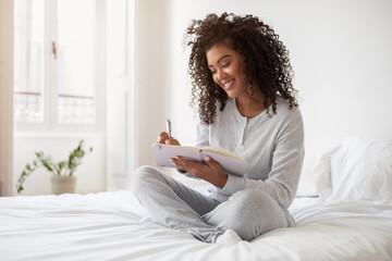 A Hispanic woman is seated on a bed, focused on writing in a notebook. She appears engrossed in her...