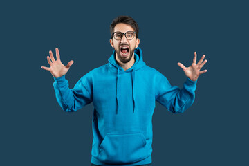 A delighted young man with glasses is standing against a dark blue backdrop, his mouth wide open as...