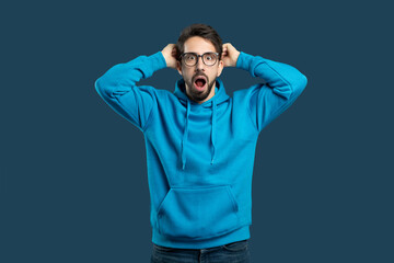 A young man wearing blue hoodie and glasses stands with his mouth open in shock, hands placed on...