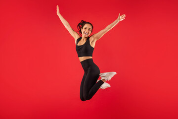 Cheerful woman wearing a black sports bra top and black leggings is captured mid-jump. She...
