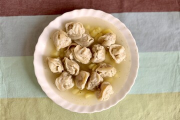 Soup with beef dumplings and potatoes in a white plate on a multi colored tablecloth on a table at home. It is steaming. Close-up