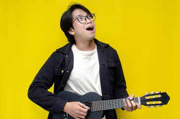 stylish ecstatic asian man playing guitar, singing, dancing and smiling isolated yellow color...
