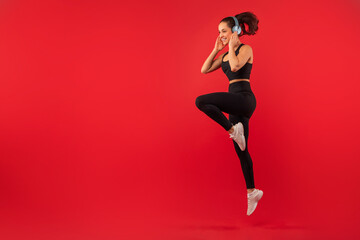 A joyful young woman with her hair tied in a ponytail is captured mid-jump against a bold red...
