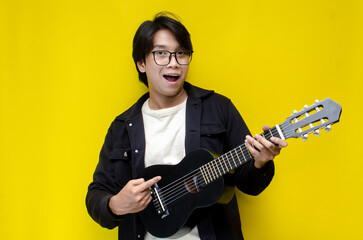 stylish ecstatic asian man playing guitar singing and smiling isolated yellow color background....