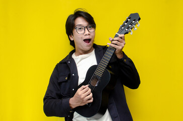 stylish ecstatic asian man playing guitar singing and smiling isolated yellow color background....