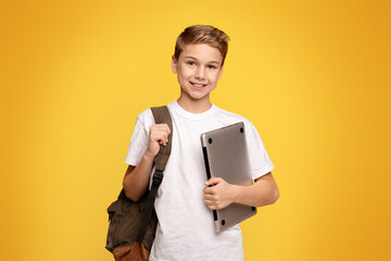 Friendly schoolboy holding laptop, going to school with computer, orange panorama background with empty space