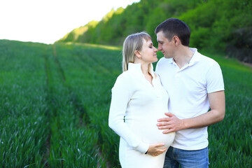 A couple standing in a beautiful field. The woman is wearing a white dress and has her hands gently placed on her stomach, indicating that she is pregnant. The man stands beside her, with his arm