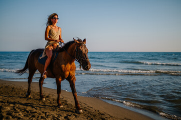 Gorgeous woman in a summer dress riding a horse on a beach during a sunset