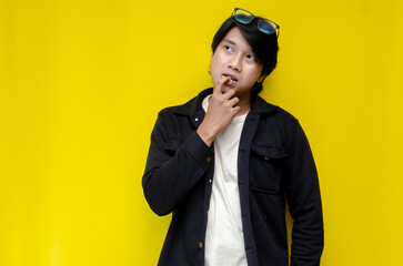 man feeling uncertain and unsure. Thoughtful asian man thinking and wondering with hand touching skin as thinking gesture isolated over yellow background.	
