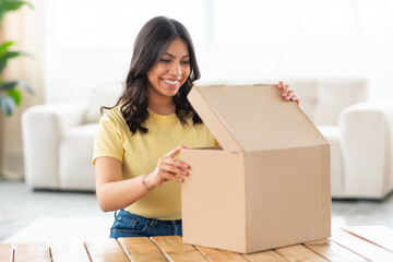 A middle eastern woman is sitting at a table, eagerly opening a box placed in front of her. She is...