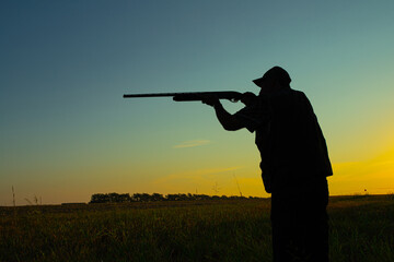 Silhouette of a hunter in a field at sunrise taking aim with a shotgun. 