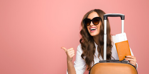 A cheerful young woman wearing large sunglasses is standing with her luggage, giving a thumbs up...