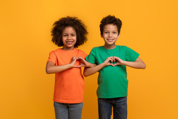 Happy African American kids boy and girl showing heart shape gestures over chest and smiling at camera, isolated on yellow studio background