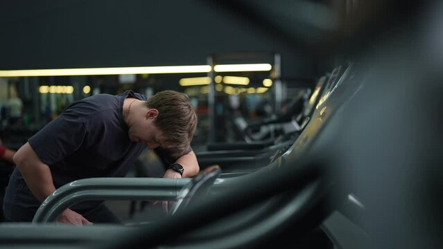 Tired beginner runner sportsman resting after running on treadmill during cardio session in gym. Satisfied fit male standing on treadmill in fitness gym, feeling exhausted after jogging training.