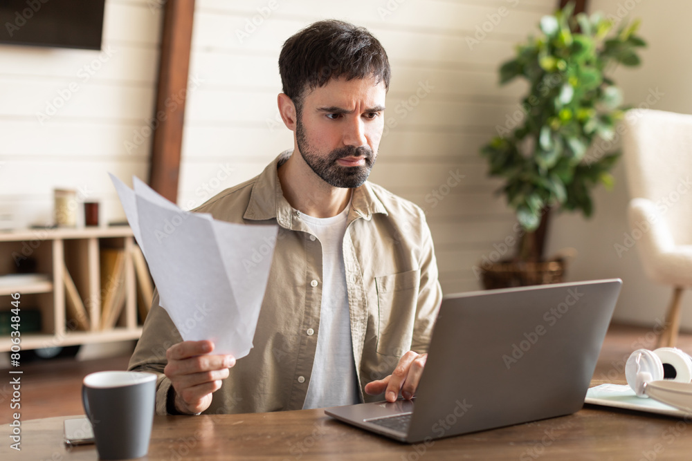Poster Distressed man is sitting at a table with a laptop open in front of him, surrounded by papers. He appears focused and engaged in his work, typing and reviewing documents.