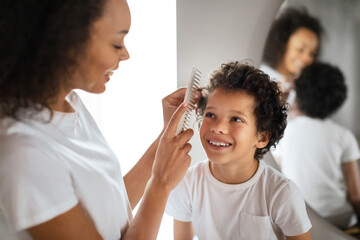 African American woman is seen combing a young boys hair in front of a mirror. Kid is sitting...