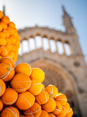 Ambiance of Fira Taronja Soller orange fair captured with the fountain adorned with a heap of ripe oranges at Plaça de la Constitució square, against the backdrop of defocused Sant Bartomeu church.