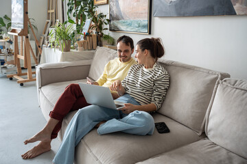 Creative relaxed happy couple sitting on comfortable couch with laptop at home art studio. Smiling...