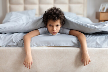 A young African American boy with curly hair is lying on his stomach, his chin resting on a bed...