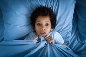 African American young boy lies in bed with a surprised look on his face, clutching the blue bed...