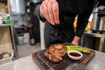 A skilled chef expertly seasons a grilled steak with asparagus and sauce on a wooden board in a professional kitchen