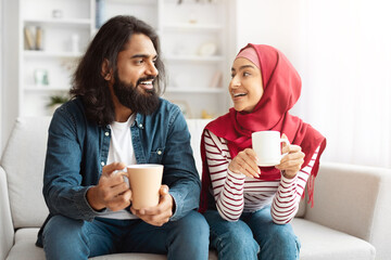 Eastern man and woman are seated comfortably on a couch, each holding a coffee cup. They appear...