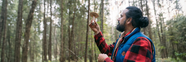 A mushroom picker man with a basket holds a mushroom in his hands in the forest.
