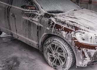 Vehicle covered in white soapy foam during regular carwash outdoors, auto getting professional wash...