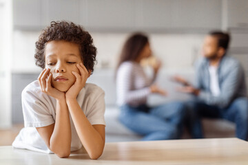 A young African American child is seated at a table, with his hands covering his face. He appears...