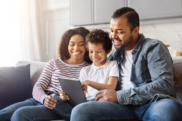 African American man, woman, and child are sitting together on a couch, engrossed in looking at a...