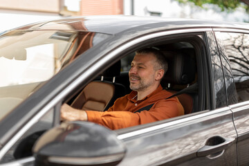 Handsome European middle aged man driving brand new car, looking at road and smiling, enjoying his...
