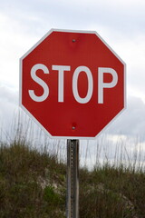 A bright red stop sign with the dunes of the beach and grey cloudy sky in the background.