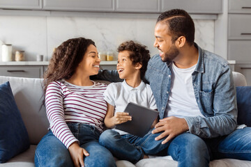 African American happy family consisting of a mother, father, and young son sits closely on a couch, sharing a moment of joy and conversation, using digital tablet
