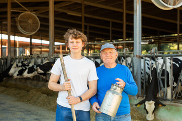 Smiling experienced elderly farmer posing with teenage grandson while working in cowshed at dairy...