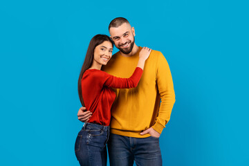 A happy couple is standing close to each other with the woman hugging the man from the side. Both are smiling and dressed in bright casual attire.
