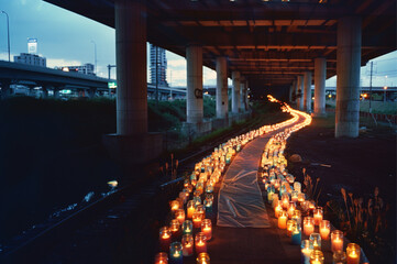 Wide view of a long, winding walking path underneath a tall highway overpass, lined with thousands of lit candles on either side, in glass jars that are covered 